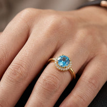 Load image into Gallery viewer, Blue oval gemstone gold ring with a halo of clear stones worn on a finger