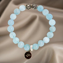 Load image into Gallery viewer, MysticFlavia Aquamarine Birthstone bracelet with light blue beads, two carved silver beads and dark brand charm, on satin fabric background