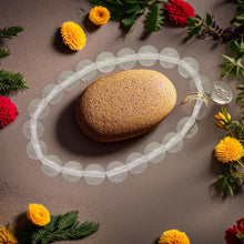 Load image into Gallery viewer, Clear quartz bracelet with silver charm displayed around a river stone, surrounded by marigolds and greenery
